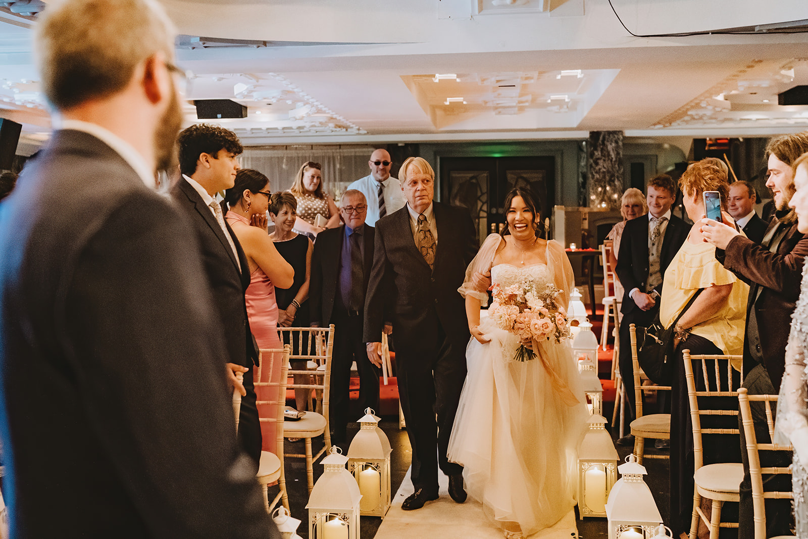 bride and father in law walking down the aisle bride has a bouquet in her hand. The grrom is waiting on the left of the photo
