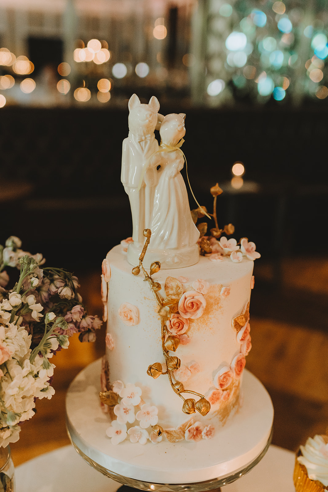 The bridal cake a tall cake with sugar flowers and a gold twig topped with two little piggies dressed as people